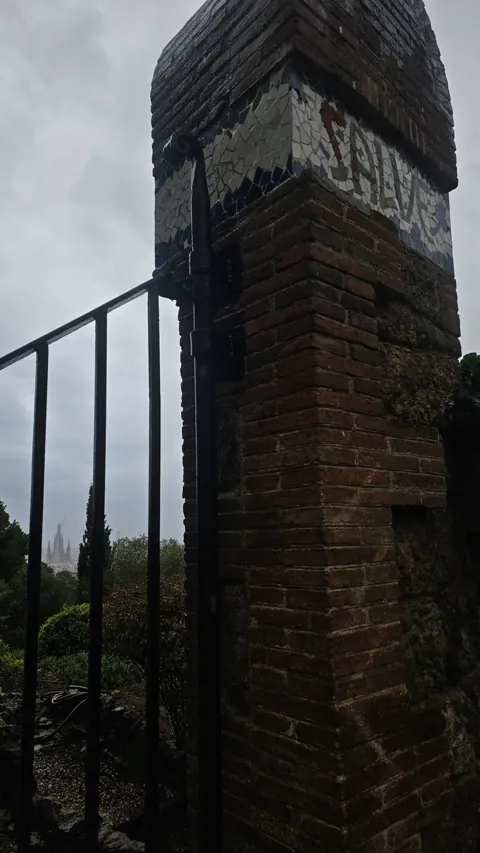 A brick and mosaic gate pillar in Barcelona against a grey sky, with an iron gate below.
