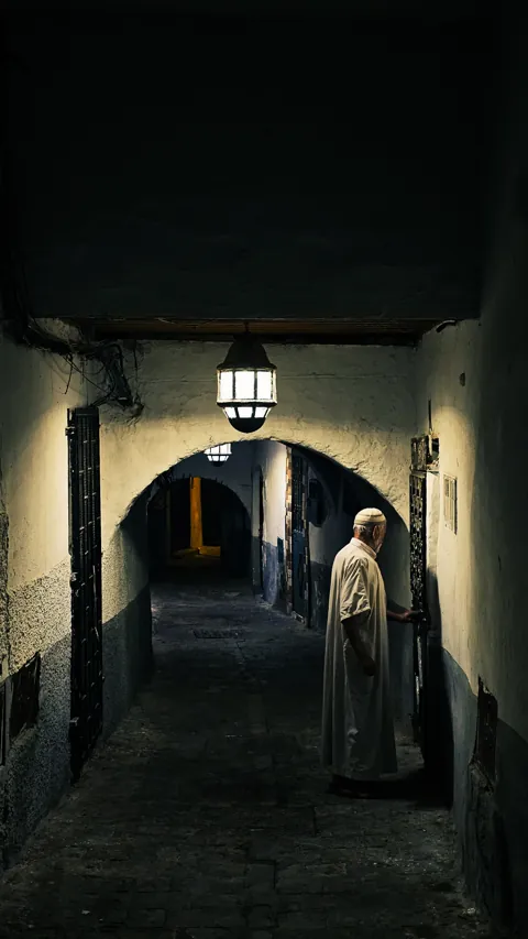 A man in a white djellaba at a doorway in a narrow alley of the Tétouan medina at night, under a hanging lantern.